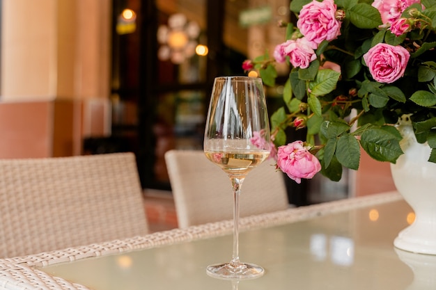 Copa de vino blanco con el telón de fondo de ramo de rosas en jarrón blanco sobre mesa de luz.