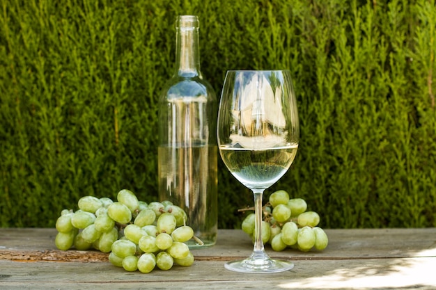 Una copa de vino blanco y uvas verdes sobre una mesa de madera con un ciprés detrás