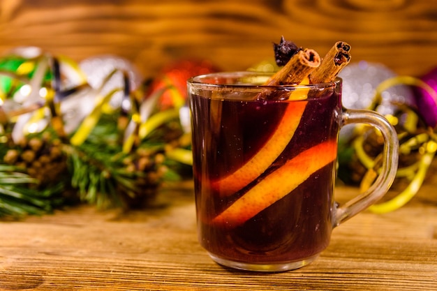 Copa de vino caliente con canela, adornos navideños y ramas de abeto en la mesa de madera rústica