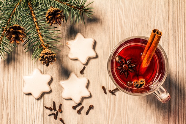 Copa de vino caliente de Navidad con canela, anís estrellado y clavo de olor sobre fondo de madera con galletas blancas, ramas de abeto natural y conos. Vista superior. Efecto tonificante de color.