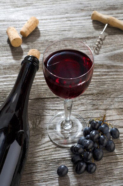 Copa de vino tinto, botella de vino, racimo de uvas azules y sacacorchos sobre una mesa de madera gris.