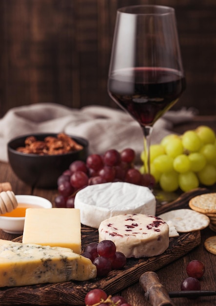Copa de vino tinto con selección de varios quesos en el tablero y uvas sobre fondo de mesa de madera. Stilton Azul, Leicester Rojo y Queso Brie.