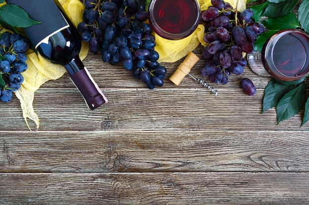 Copas de vino con vino tinto, botella, sacacorchos, uvas azules, hojas sobre una mesa de madera.