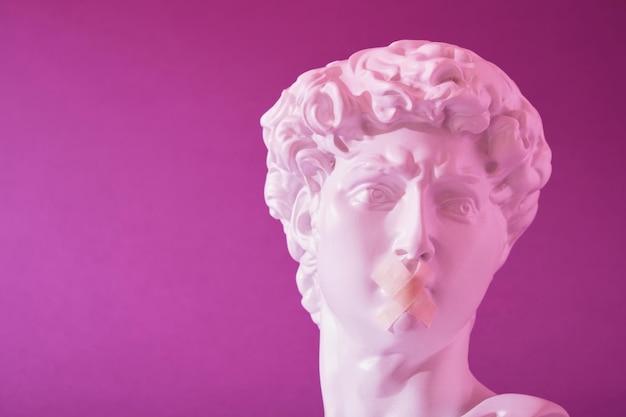 Copia de la cabeza de una estatua antigua de David con una boca tapada con luz de neón rosa sobre un fondo violeta