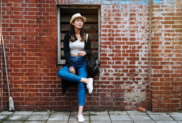 Foto copie el espacio retrato completo de una joven coreana asiática con camisa corta y jeans azules de pie contra la pared de ladrillo rojo. dama relajada con sombrero de paja disfruta de las vacaciones de verano y viaja con mochila