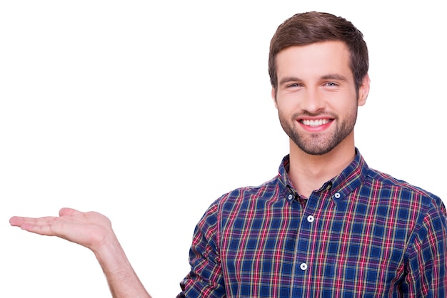 Copie el espacio en su mano. Retrato de hombre joven guapo en camisa casual con espacio de copia y mirando a la cámara con una sonrisa mientras está de pie aislado en blanco