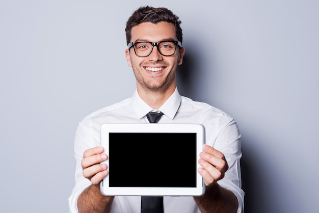 Copie el espacio en su tableta. Apuesto joven en camisa y corbata mostrando su tableta digital y sonriendo mientras está de pie contra el fondo gris