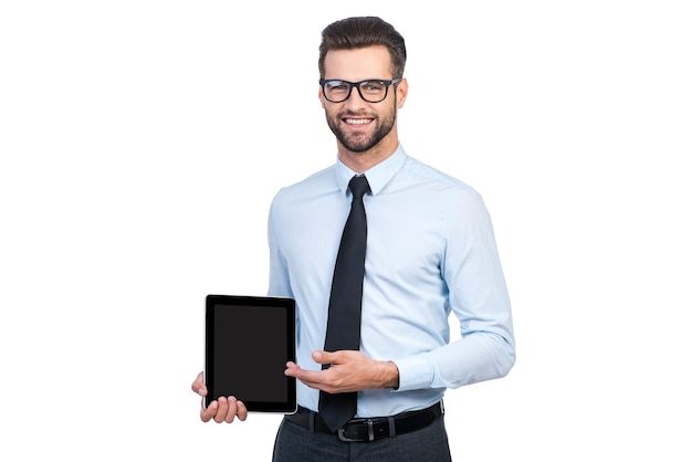 Copie el espacio en su tableta. Hombre guapo joven confiado en camisa y corbata sosteniendo tableta digital y apuntando con una sonrisa mientras está de pie contra el fondo blanco