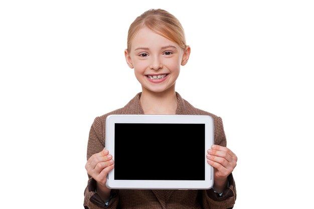 Copie el espacio en su tableta. Niña alegre en ropa formal mostrando su tableta digital y sonriendo mientras está de pie aislado en blanco