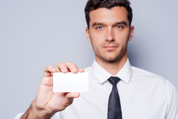 Copie el espacio en su tarjeta de presentación. Hombre joven confidente en camisa y corbata mostrando su tarjeta de visita mientras está de pie contra el fondo gris