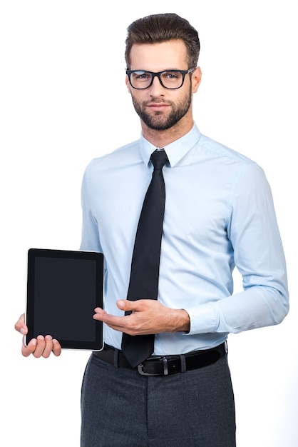 Copie o espaço em seu tablet. Homem bonito jovem confiante de camisa e gravata segurando um tablet digital e apontando-o com um sorriso em pé contra um fundo branco