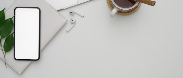 Copie o espaço na mesa de trabalho branca com smartphone de tela em branco, fones de ouvido sem fio, agenda e xícara de chocolate quente