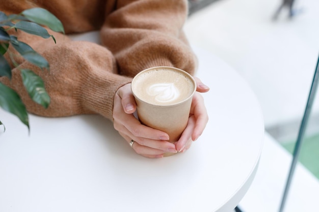 Copo artesanal com cappuccino nas mãos de uma garota em close-up da mesa