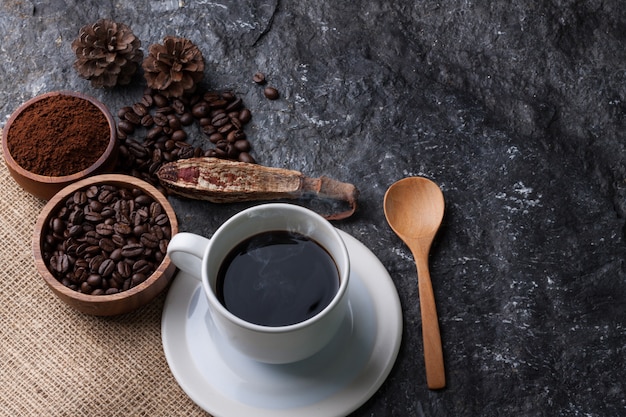 Copo branco café, grãos de café em copo de madeira na serapilheira, colher de madeira no fundo de pedra preto