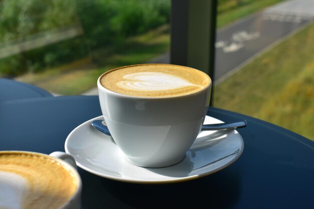 Copo branco com cappuccino e espuma em forma de coração em uma mesa azul perto de uma janela panorâmica