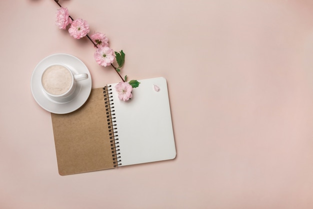Copo branco com cappuccino, flores de sakura, caderno em um fundo rosa pastel