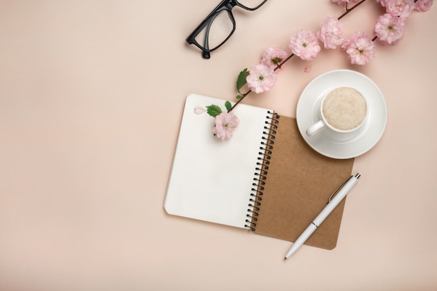 Copo branco com cappuccino, flores de sakura, caderno em um fundo rosa pastel