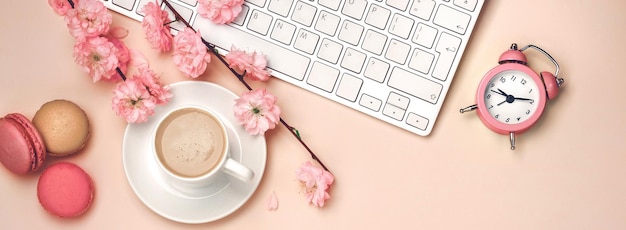 Copo branco com cappuccino, flores de sakura, teclado, macarons, despertador em um fundo rosa pastel. vista do topo.