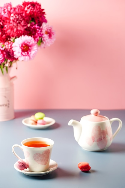 Copo branco com corações rosa um bule, doces e um buquê de flores em um fundo cinza-rosa. Café da manhã, chá da manhã.