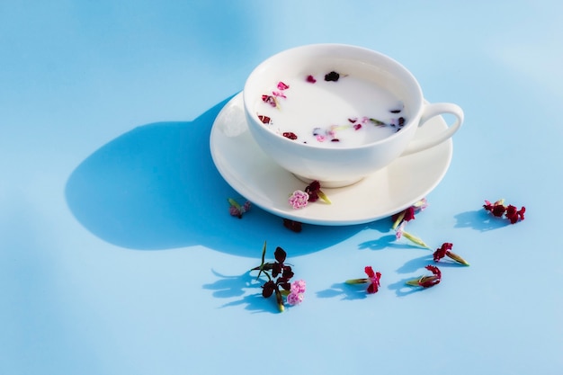 Copo branco com leite vegan e flores sobre fundo azul. Conceito de bebidas e alimentos vegetarianos, chás de ervas, beleza e saúde, spa.