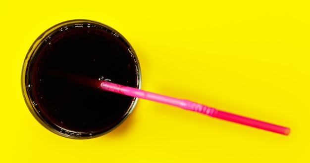 Copo com bebida de cola e palha rosa única na mesa amarela, vista de cima