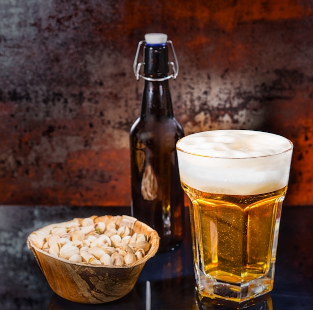 Copo com cerveja light derramada na hora, garrafa de cerveja perto da placa de madeira com pistache em uma superfície de espelho preto. Conceito de alimentos e bebidas