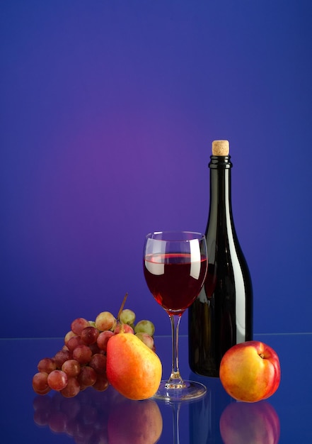 Copo com vinho tinto, garrafa de vinho, escova de uvas e frutas no fundo multicolorido brilhante com reflexão. cópia-espaço.