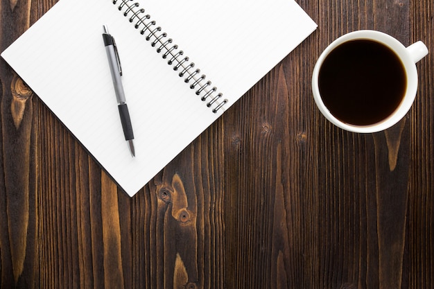 Copo de café branco com café preto, caderno e pena na tabela de madeira.