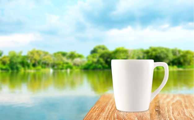 Copo de café branco na mesa de madeira em árvore borrada e fundo da lagoa