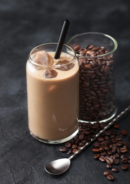 Copo de café gelado e leite com grãos de café crus grátis em recipiente de vidro com colher longa sobre fundo preto.