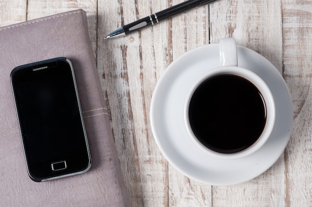 Copo de café quente, telefone e caderno em madeira branca