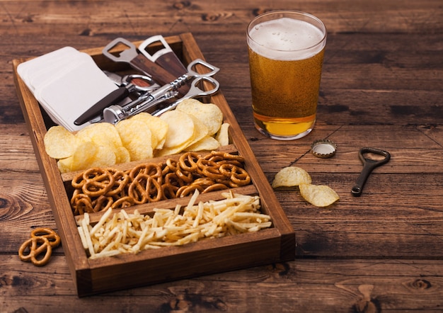 Copo de cerveja artesanal e abridor com caixa de lanches em fundo de madeira. Pretzel, batatas fritas e palitos de batata salgada em caixa de madeira vintage com abridores e tapetes de cerveja.