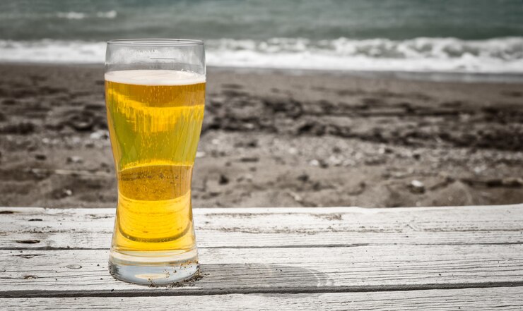 Foto copo de cerveja pilsner dourada clara em uma mesa rústica ao ar livre na areia à beira-mar com areia escura e ondas quebrando ao fundo
