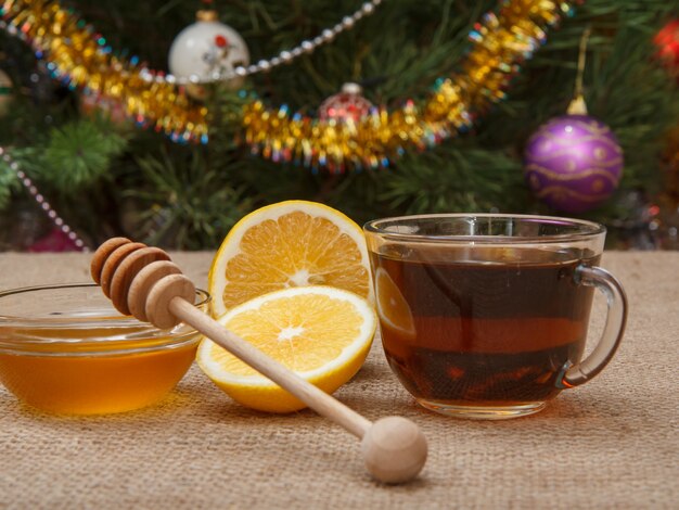 Copo de chá de close-up, pedaços de limão e mel em uma tigela de vidro com uma colher de pau na mesa com serapilheira e a árvore de natal com bolas de brinquedo e guirlandas no fundo.