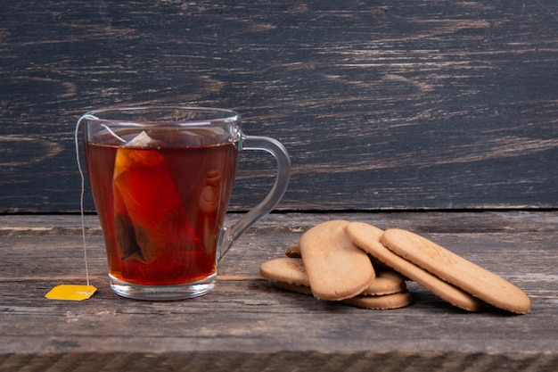 Copo de chá preto com saquinho de chá e biscoito em uma mesa de madeira