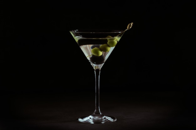 Copo de cocktail de martini seco clássico com azeitonas na mesa de pedra escura contra um negro.