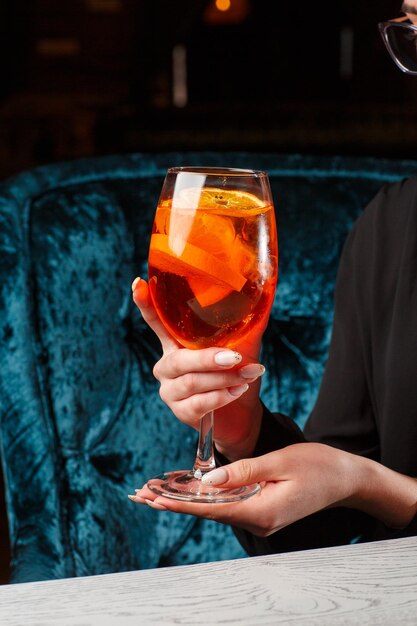 Copo de coquetel Aperol spritz gelado servido em um copo de vinho nas mãos femininas decorado com fatias de laranja colocadas em um fundo preto