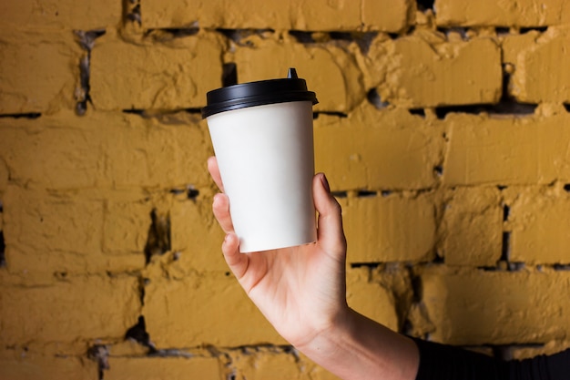Copo de papel branco com café na mão contra uma parede de tijolos amarelos