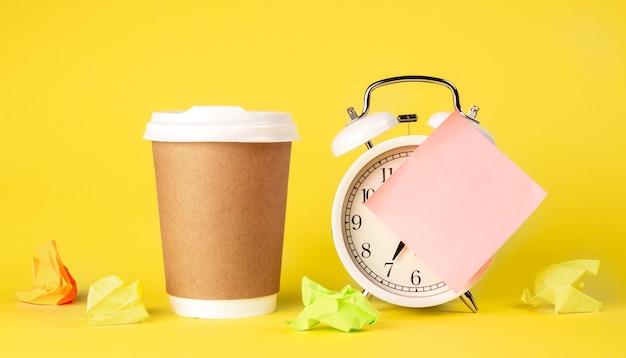 Copo de papel e despertador com lembrete de papel no fundo amarelo isolado