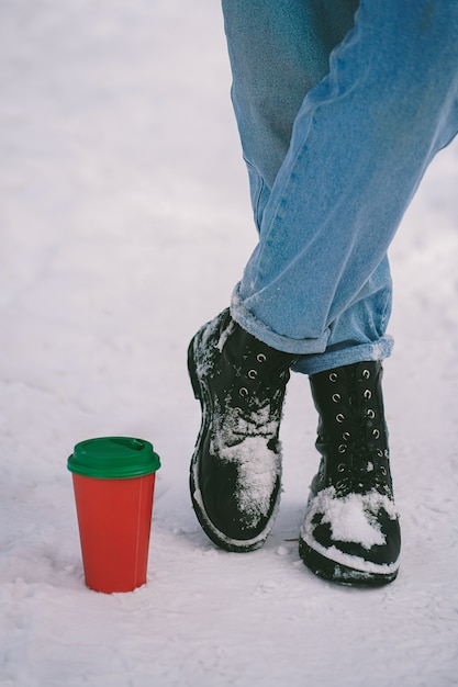 Foto copo de papel fica na neve próximo a pés em botas