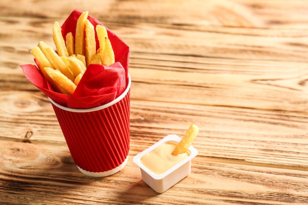 Copo de papel vermelho cheio de deliciosas batatas fritas com molho de queijo no fundo de madeira