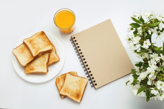 Copo de suco de laranja, brindes em um prato branco, bloco de notas e um ramo com flores sobre fundo branco. Conceito de um saudável café da manhã de primavera