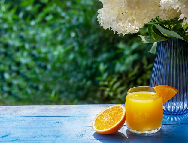 Copo de suco de laranja fresco decorado com uma fatia de laranja madura na mesa de madeira azul com flores no fundo