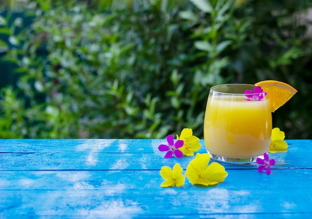 Copo de suco de laranja fresco guarnecido com uma fatia de laranja madura e flores na mesa de madeira azul