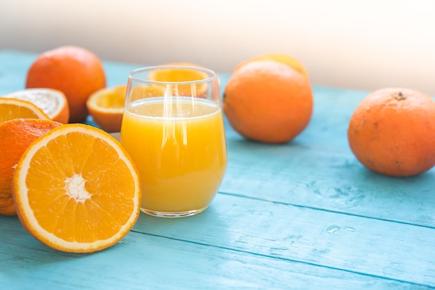 Copo de suco natural de várias laranjas orgânicas, algumas inteiras, outras espremidas, para a dieta, sobre uma mesa de madeira azul. Café da manhã saudável