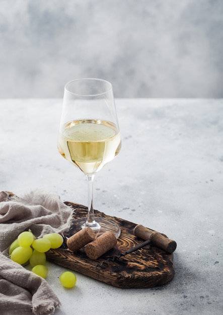 Copo de vinho caseiro branco com rolhas, saca-rolhas e uvas na placa de madeira com pano de linho no fundo da mesa de luz.