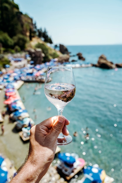 Copo de vinho na mão com vinho branco fresco jovem no fundo da praia mediterrânea ...