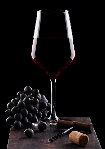 Copo de vinho tinto com uvas escuras e abridor de saca-rolhas vintage e cortiça na placa de madeira no preto