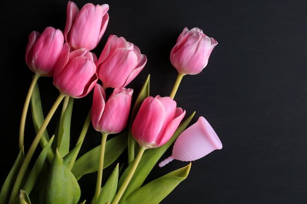 Copo menstrual e tulipas cor de rosa em um fundo preto. Saúde da mulher.
