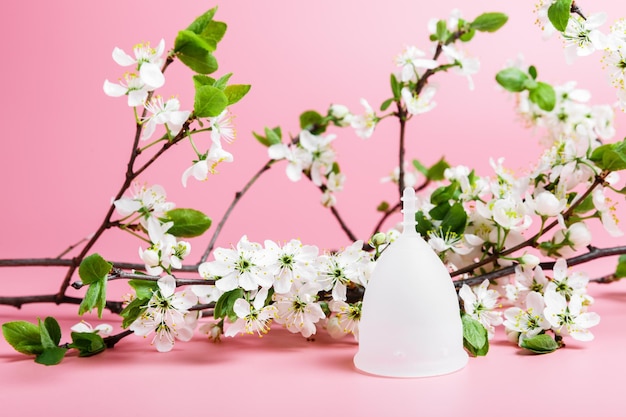 Copo menstrual eco branco reutilizável em um fundo rosa galhos de cerejeira de primavera com flores brancas conceito de ecologia e reciclagem desperdício zero menstruação de higiene feminina dias críticos